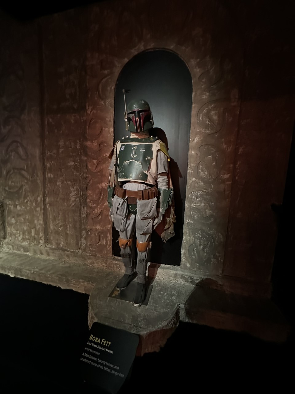 Boba Fett's weathered Mandalorian armor with jetpack and weaponry displayed in stone alcove