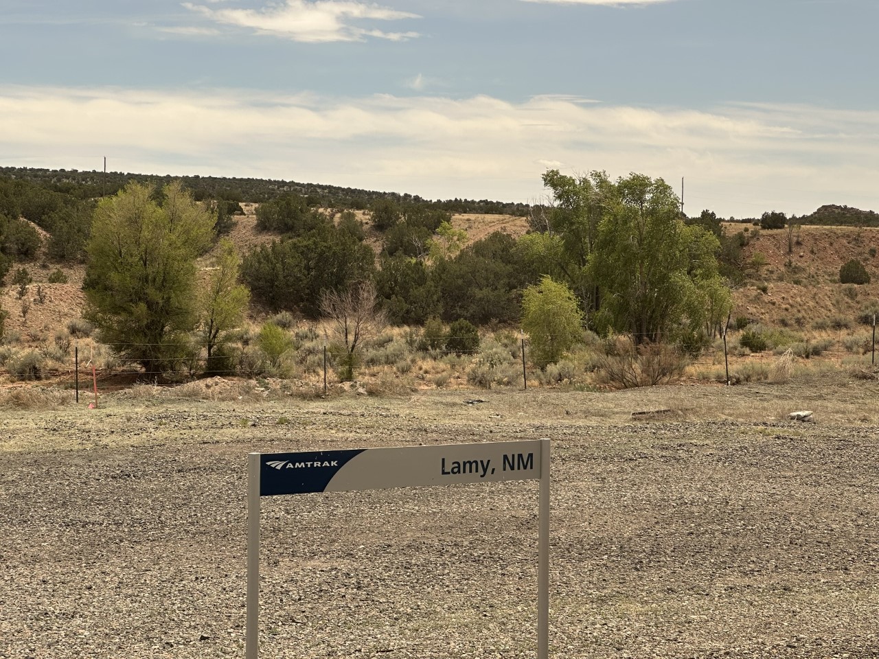 Amtrak 'Lamy, NM' platform sign in a gravel lot, juniper-dotted pink hills behind the Santa Fe stop