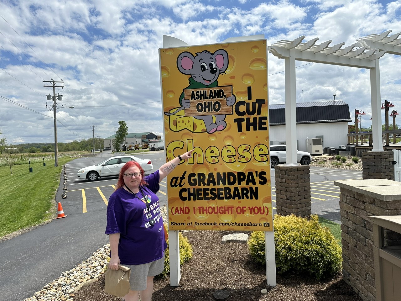 Rachel pointing at the large yellow I Cut the Cheese at Grandpa's Cheesebarn photo-op sign in the parking lot
